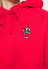 Sweat à capuche rouge avec cordon de serrage, design brodé du personnage Mario, et une petite étiquette logo "Levi's" sur le devant.
