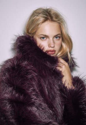 Blonde vrouw met blauwe ogen die een grote donkerpaarse nepbontjas draagt, terwijl ze de kraag dicht tegen haar gezicht houdt voor een effen achtergrond.
