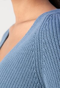 Gros plan du cou et de l'épaule d'une personne portant un pull tricoté bleu clair avec une texture côtelée.