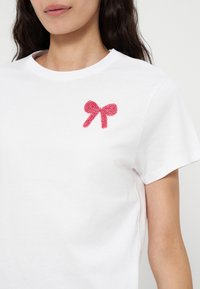 Femme portant un t-shirt blanc avec un petit nœud rouge en perles sur la partie supérieure gauche de la poitrine, manches courtes et col rond.