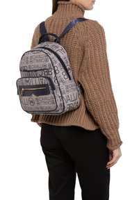 Zaino caratterizzato da un tessuto testurizzato con un motivo logo ripetuto in blu navy e beige, doppi scomparti con zip e spallacci regolabili.