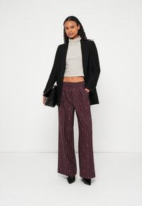 Zwarte blazer over een gestreepte, aansluitende coltrui en wijde, bordeauxrode broek met een verticale streepjespatroon. Zwarte handtas en puntige schoenen.