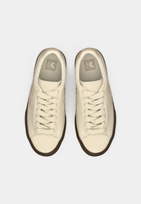 Sneakers in tela beige con punta rotonda, lacci piatti e suole in gomma marrone. Presentano il logo a forma di "V" sui lati e sulle solette.