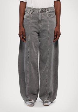 Person iført vidde-grå jeans og sølvfarvede spidse sko, stående med hænder afslappet ved siden.