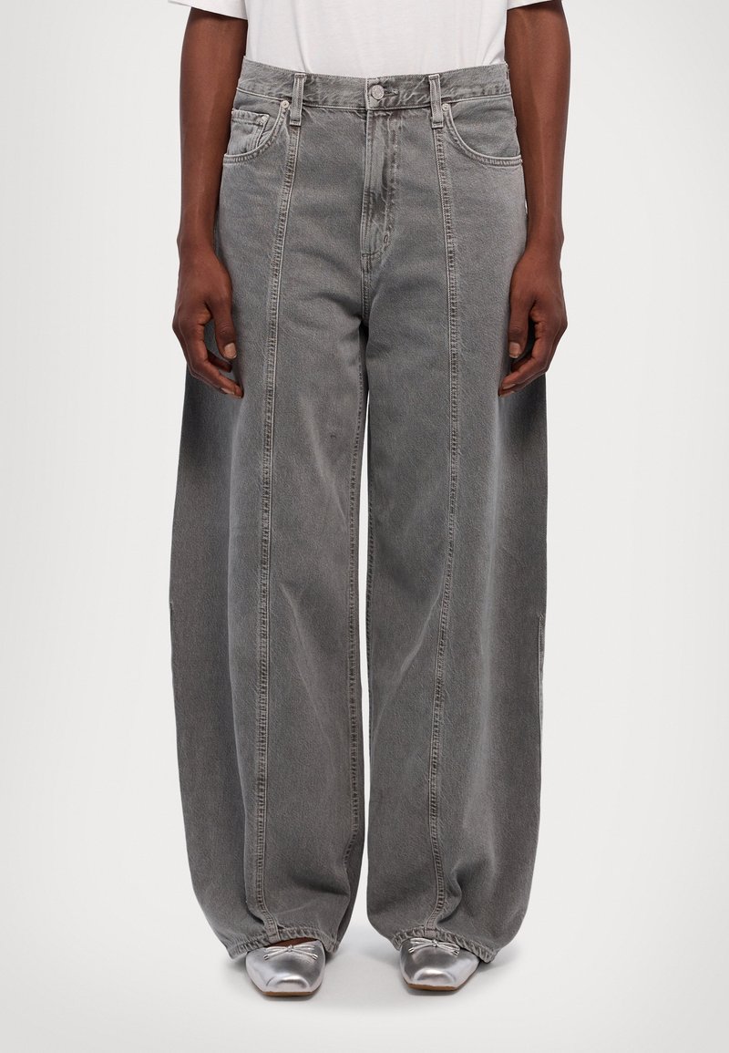 Personne portant un jean gris à jambes larges et des chaussures argentées à bout pointu, debout avec les mains détendues le long du corps.