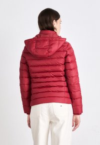 Červená páperová bunda s horizontálne prešívanými panelmi, kapucňou a elastickými manžetami. Tkanina má hladkú textúru a priliehavý strih.