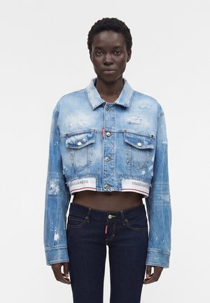 Vrouw die een cropped, lichtblauw distressed spijkerjasje en donkere high-waisted jeans draagt, staand tegen een effen witte achtergrond.