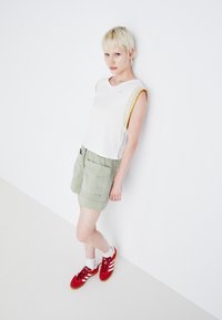Hvit ermeløs topp med gule og beige bølgekantede detaljer, kombinert med lysegrønne shorts og røde joggesko med hvite striper.