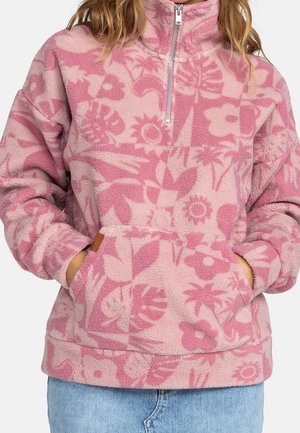 Vrouw draagt een roze fleece pullover met bloemen- en bladmotieven, voorrits en voorzak, gecombineerd met een blauwe jeans.