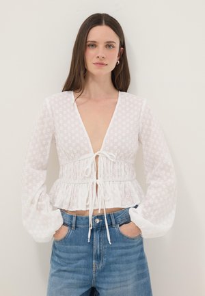 Donna con capelli lunghi castani che indossa una blusa floreale bianca trasparente con maniche a sbuffo e jeans blu, con le mani nelle tasche, su sfondo semplice.