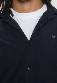 Camicia di velluto a coste blu navy con bottoni e colletto appuntito, con una t-shirt bianca girocollo sotto e un logo ricamato discreto sul petto.