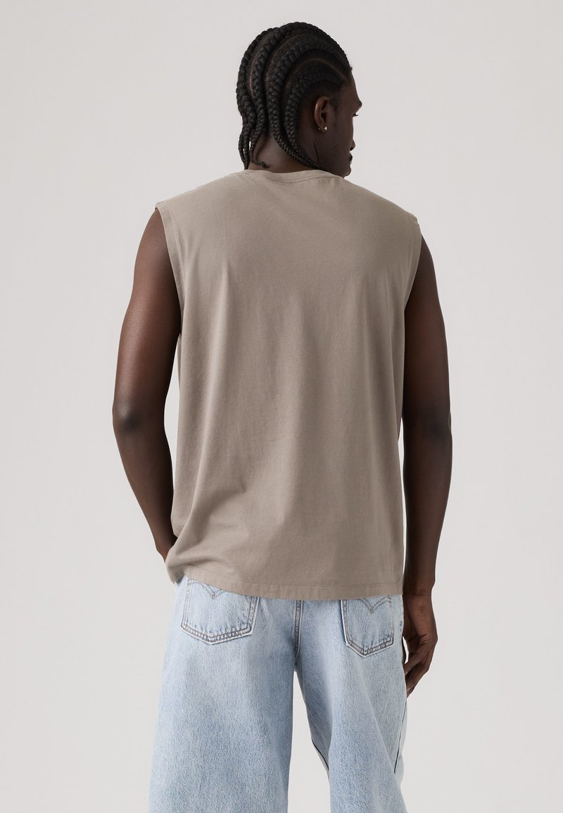 Mann mit geflochtenem Haar, der ein ärmelloses beiges Hemd und hellblaue Jeansshorts trägt, steht mit dem Rücken zu einem einfarbigen Hintergrund.
