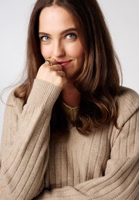 Pull beige côtelé à manches longues, mettant en valeur un tricot texturé avec une légère brillance, accompagné d'un collier statement doré et de plusieurs bagues.