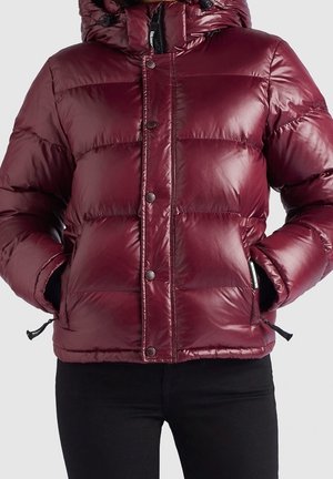 Doudoune bordeaux brillante avec boutons-pression, poches zippées et capuche, portée sur un pantalon noir avec les mains dans les poches.