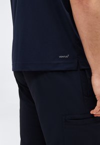 Άτομο που φορά μπλε σκούρο πουκάμισο με το λογότυπο «VENTUS» κοντά στο τελείωμα και αντίστοιχο μπλε cargo παντελόνι με πλαϊνή τσέπη, με το χέρι να ακουμπάει στο μηρό.