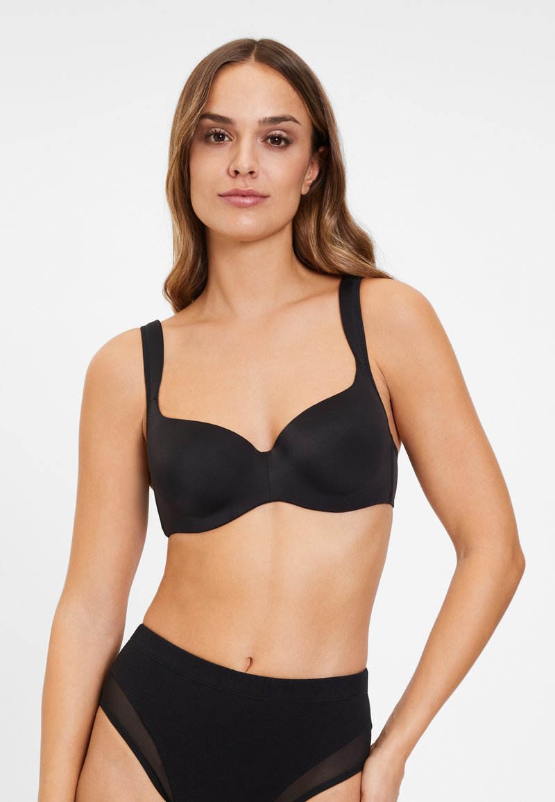 Sostén negro con aros de alambre, fabricado en un tejido mate suave, con copas contorneadas y tirantes anchos ajustables. Combinado con bragas negras de talle alto que presentan detalles de malla.