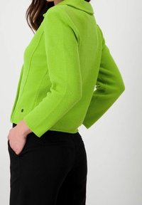 Chaqueta texturizada verde lima con cuello, diseño ajustado y mangas largas, combinada con pantalones negros. Detalle de botones notable en el dobladillo.