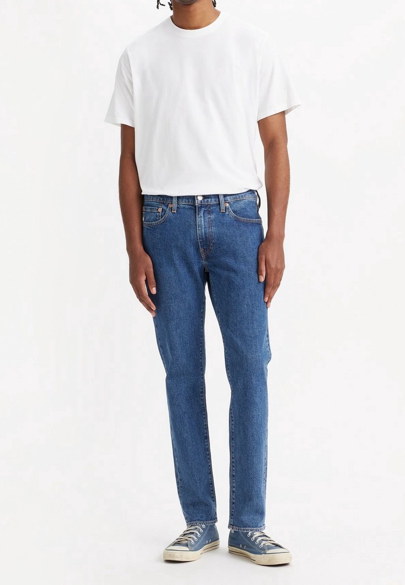 T-shirt blanc à col rond, jean en denim bleu et baskets en toile bleue. Le denim a une coupe classique avec des coutures visibles et des détails métalliques.