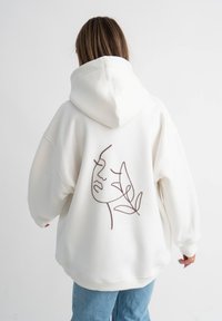 Felpa bianca con un grande design lineare marrone di un volto e una foglia sul retro. Ha una vestibilità ampia e rilassata, oltre a un cappuccio.