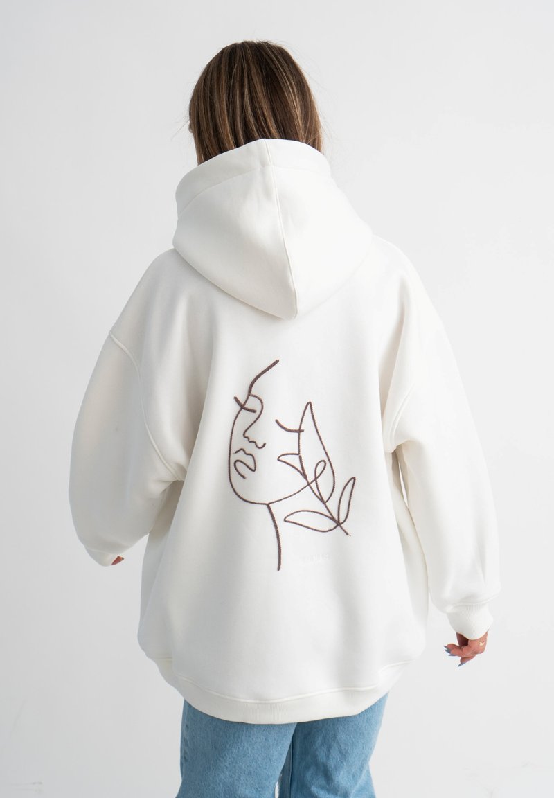 Felpa bianca con un grande design lineare marrone di un volto e una foglia sul retro. Ha una vestibilità ampia e rilassata, oltre a un cappuccio.