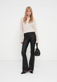 Pantalons noirs évasés avec une finition brillante, associés à un haut à manches longues beige clair et un petit sac à main noir texturé.