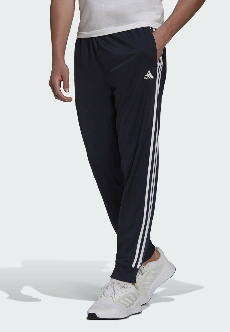 Marineblaue Sporthose mit weißem Drei-Streifen-Design, elastischem Bund, schmal zulaufender Passform und Seitentaschen, kombiniert mit weißen Laufschuhen.