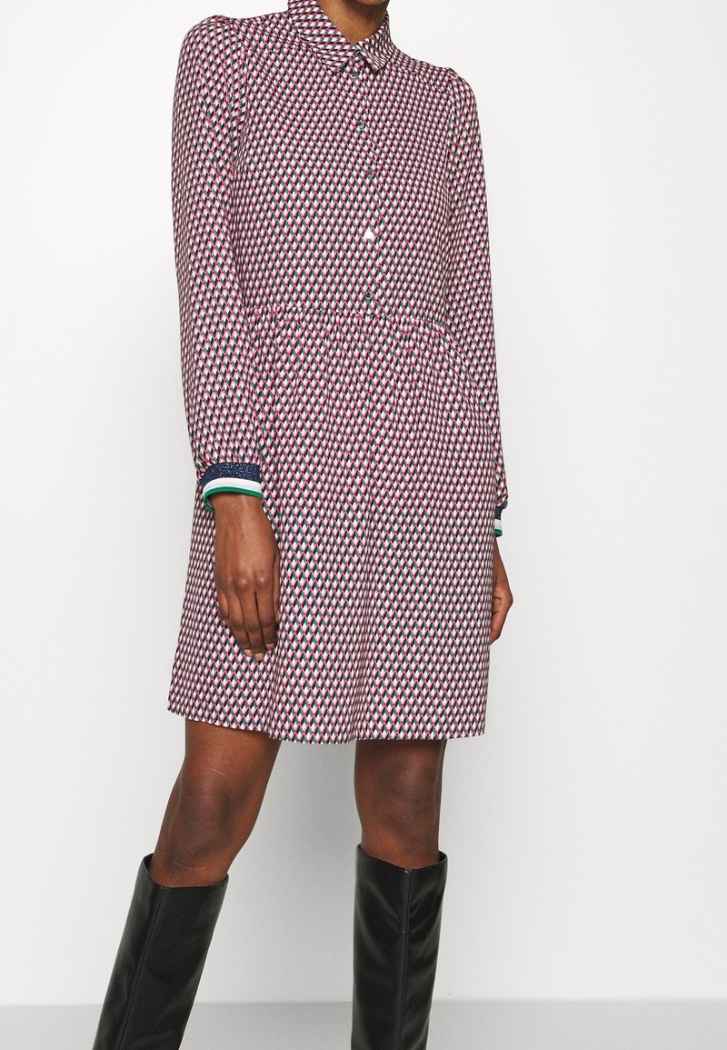 Femme portant une robe mi-longue à manches longues avec un motif géométrique rose, noir et blanc, boutonnée sur le devant, et des bottes noires montant jusqu'aux genoux.