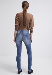 Jeans ajustados de color azul con un lavado desgastado y un ligero desgaste, combinados con una camiseta de manga larga marrón, vista desde la parte de atrás.