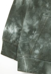 Primer plano de tela de tie-dye verde con tejido texturizado, puños acanalados y costuras de bolsillo en ángulo en una prenda.