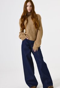 Hellbrauner, gestrickter Pullover mit lockerer Passform und langen Ärmeln, kombiniert mit dunkelblauen, weiten Jeans. Weiche Textur, lässiges Design.