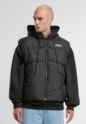 Črn puffer brezrokavnik s kapuco, quiltana tekstura, sprednji zadrga in značkasta nalepka na prsih, nosi pa ga nad črno majico z dolgimi rokavi.