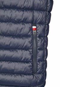 Marineblauer Steppweste mit genähtem Design, ausgestattet mit einer seitlichen Reißverschlusstasche, die von einem rot-weiß-blauen Logo-Besatz akzentuiert wird. Glatte Textur.