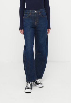 Personne portant un jean large taille haute bleu foncé associé à des baskets noires et blanches et un haut à manches longues bleu marine.
