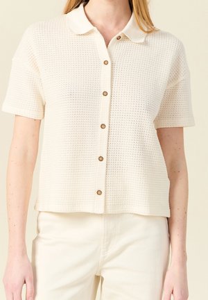 Femme portant une chemise en tricot crème à manches courtes avec col et boutons, associée à un pantalon taille haute crème, sur un fond neutre.