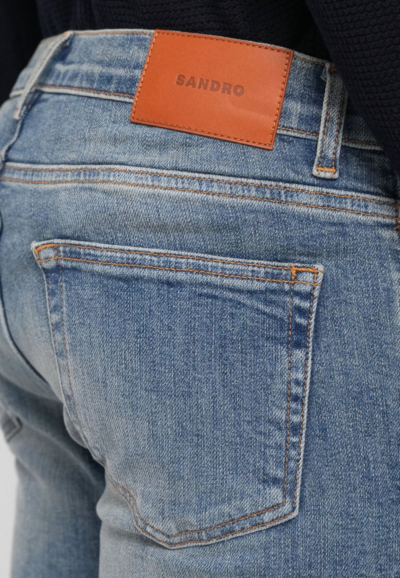 Lys blå denim jeans med et brunt lædermærke med teksten "SANDRO" på den bageste talje. Har kontrasterende orange syninger og traditionelle lommer.