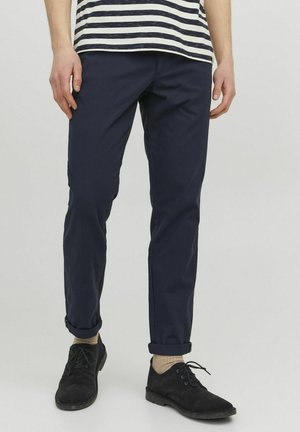 Pantaloni chino blu navy dal taglio slim, con risvolti e tessuto liscio e traspirante. Scarpe nere in camoscio e calzini di colore chiaro completano il look.