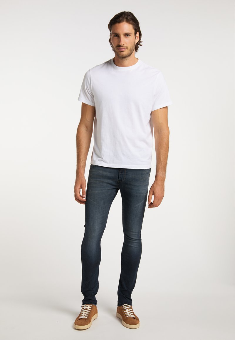 Mann, der vor einem einfarbigen Hintergrund steht, trägt ein weißes T-Shirt, dunkelblaue Skinny-Jeans und braune Sneakers mit weißen Schnürsenkeln.