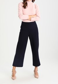 Pantalon large bleu marine à la texture lisse. Associé à un col roulé côtelé rose clair et des talons beiges à lanières. Design simple et moderne.