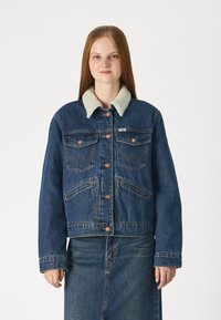 Τζάκετ από τζιν με ελαφρύ, αφράτο γιακά. Διαθέτει δύο τσέπες στο στήθος, μπροστινό κούμπωμα με κουμπιά και αντίθετη ραφή. Σκούρο μπλε ύφασμα.