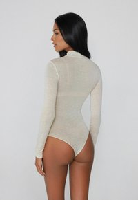 Bodysuit z długim rękawem w jasnym beżu, wykonany z przezroczystego materiału. Posiada stójkę i projekt z tyłu w stylu stringów, podkreślający kontur ciała.