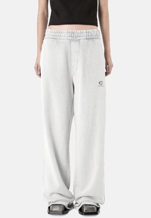 Persona che indossa pantaloni larghi grigio chiaro con vita elasticizzata, scarpe nere dalla punta quadrata e un top corto nero su uno sfondo semplice.