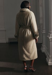 Beige wollen jas met een ceintuur in de taille, grote kraag en gestructureerde stof. Halflang tot mid-kuit met opgerolde manchetten, gecombineerd met sandaalhakken.