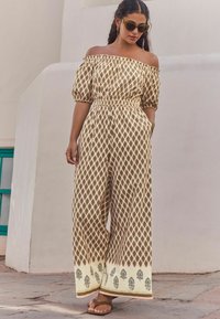 Off-shoulder jumpsuit i krämfärg med ett brunt geometriskt mönster, elastisk midja, vida ben och dekorativa detaljer i nederkant.