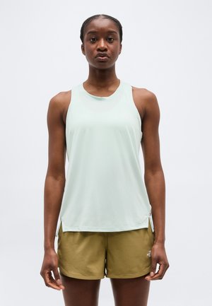 Žena stojaca čelom dopredu, oblečená v svetlomodrom bezrukávovom topu a olivovo zelených krátkych nohaviciach na jednoduchom pozadí.