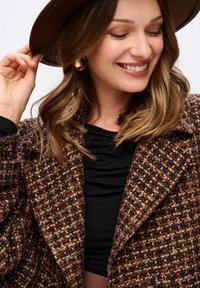 Brauner Tweed-Mantel mit strukturiertem Muster, schwarzes Oberteil darunter und ein breitkrempiger Hut; goldene Creolen ergänzen den Look.
