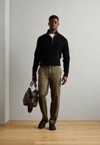 Pull noir à côtes avec une fermeture éclair quart, pantalon vert olive, portant un sac vert foncé et des chaussures noires sur un sol en bois.
