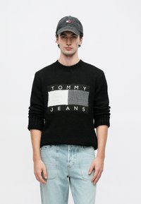 Μαύρο πλεκτό πουλόβερ με την επιγραφή "TOMMY JEANS" σε λευκό κείμενο και γκρι-λευκά πάνελ με χρωματικές αντιθέσεις. Ανοιχτόχρωμο μπλε τζιν και γκρι καπέλο.