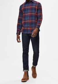 Mansrutig skjorta i marinblå, röd och grå nyanser, kombinerad med mörka jeans och bruna mockakängor. Skjortan har lång ärm och knappar.