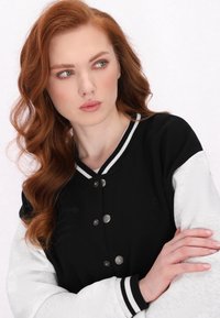 Jachetă varsity alb-negru, cu nasturi tip snap, mâneci contrastante, guler ribat și broderie subtilă în zona pieptului.