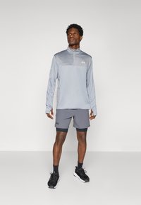 Svijetlo siva dugih rukava sportska majica s ovratnikom na zip, u kombinaciji s tamnosivim šortsevima. Oba komada imaju logo Under Armour. Crne sportske tenisice.